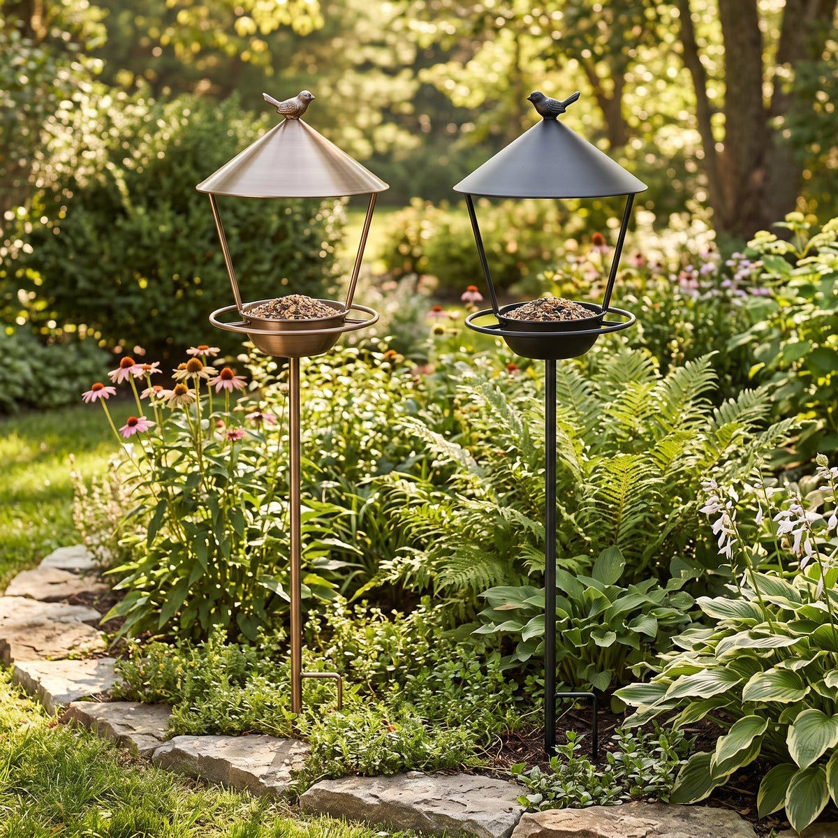 Brushed Bronze and Matte Black standing metal bird feeding stations staked side by side in a blooming cottage flower garden
