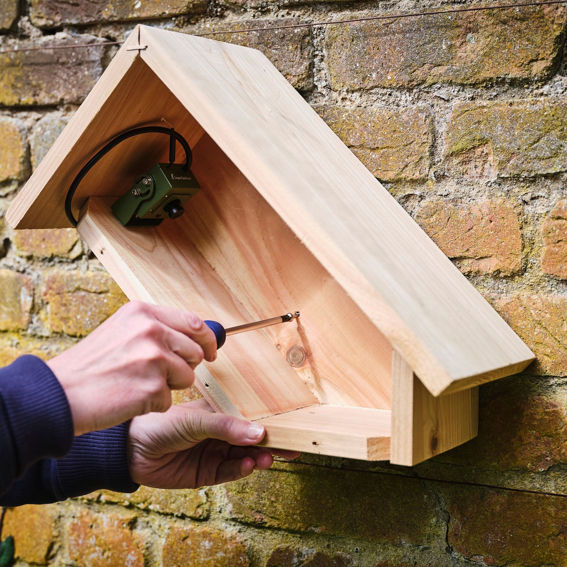 Person assembling a wooden birdhouse against a brick wall