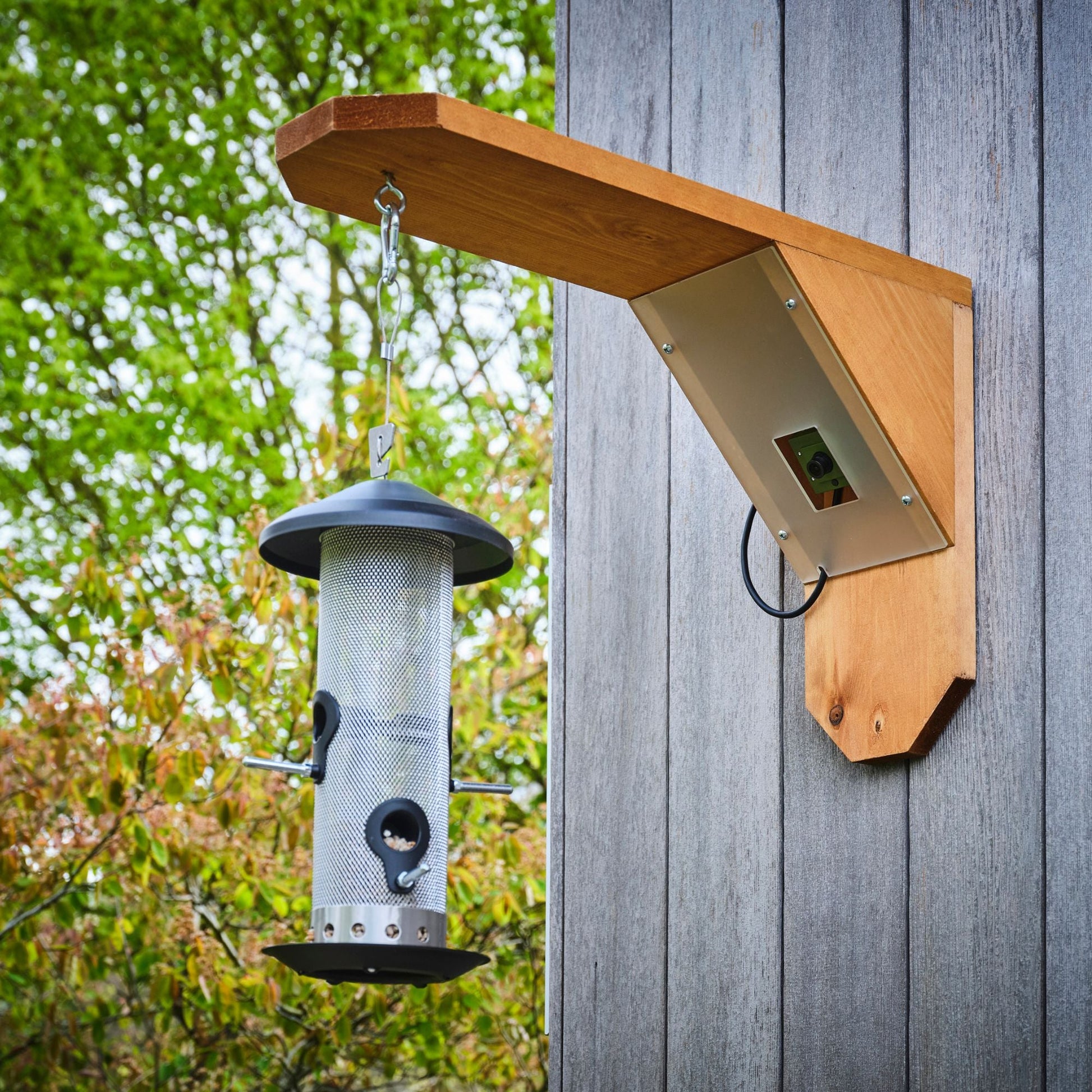 Bird feeder hanging from a wooden bracket on a wooden wall with trees in the background