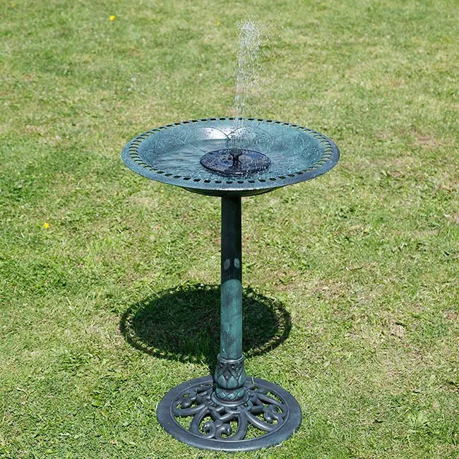 A green metal pedestal bird bath with a small central fountain spraying water, set on a green grass lawn.