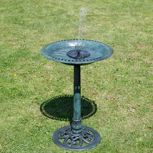 A green metal pedestal bird bath with a small central fountain spraying water, set on a green grass lawn.