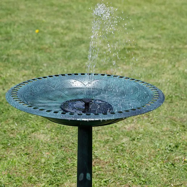 A round black metal bird bath fountain with water spraying upward, set on a green grass lawn.