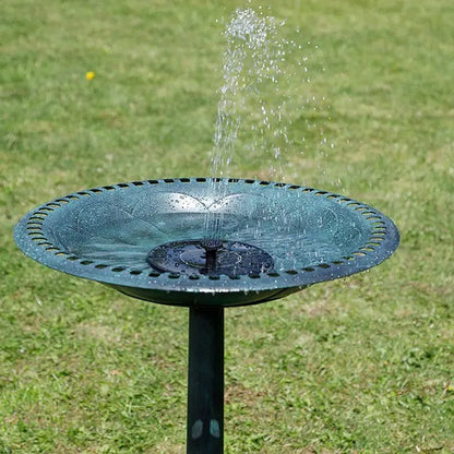 A round black metal bird bath fountain with water spraying upward, set on a green grass lawn.